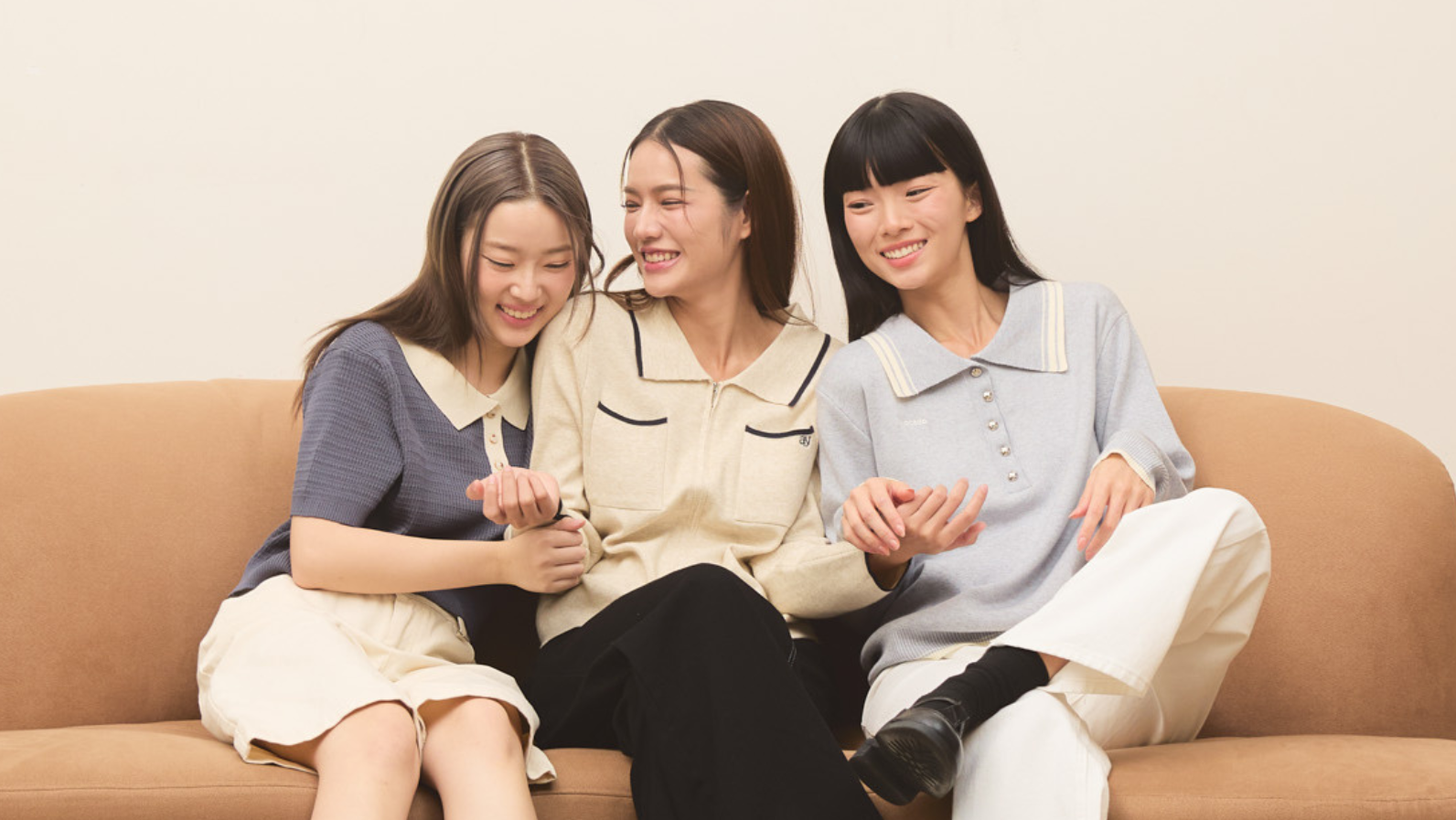 Three women sitting together on a tan couch against a plain beige wall, wearing coordinated casual outfits in blue, cream, black, and white.